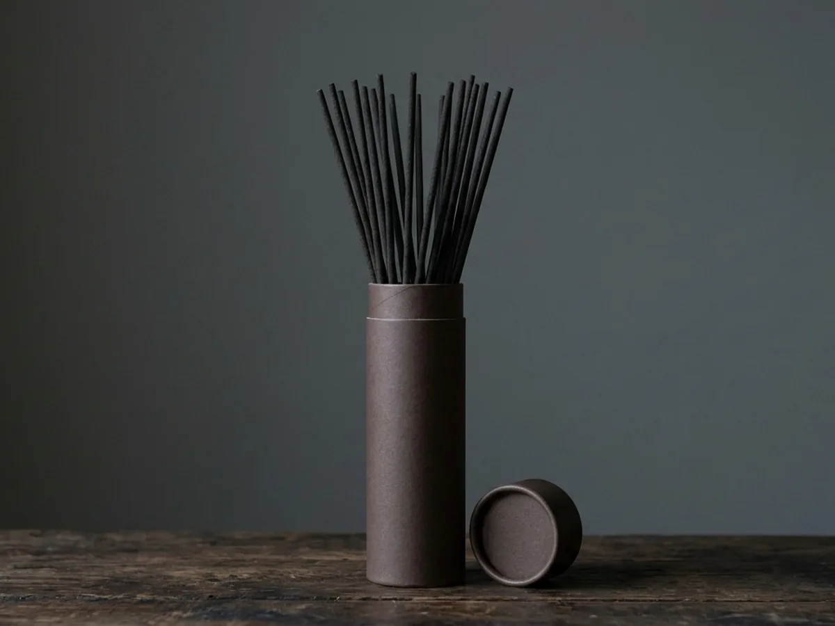 Plain kraft tube box standing open on dark timber, long dark oud incense sticks emerging from the top