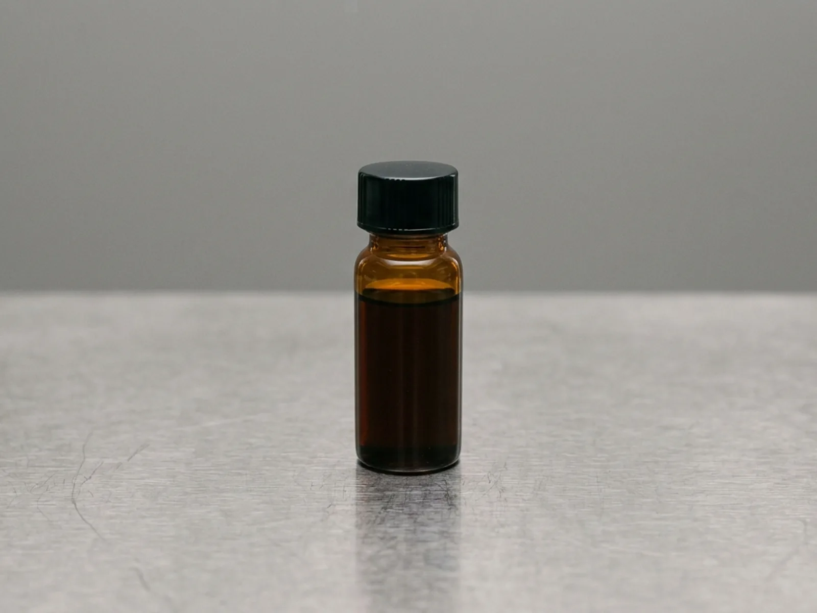 A single unlabelled amber vial of dark oil standing alone on a stainless bench with nothing else present