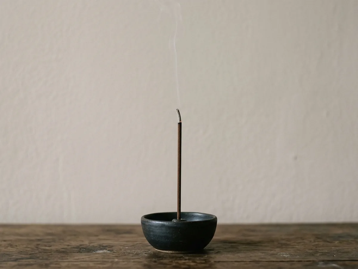 A single coreless agarwood incense stick burning upright in a small dark ceramic holder, one thin thread of smoke