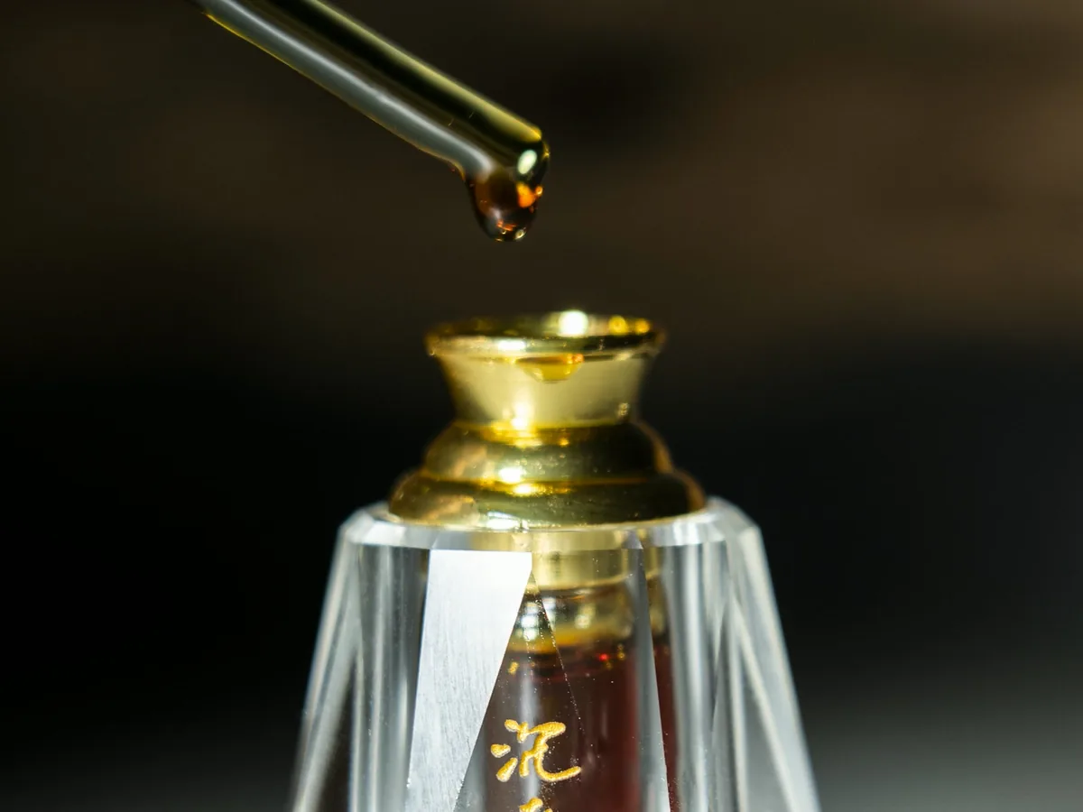 A drop of dark oud oil hanging from a glass pipette above the open neck of a crystal tola flask