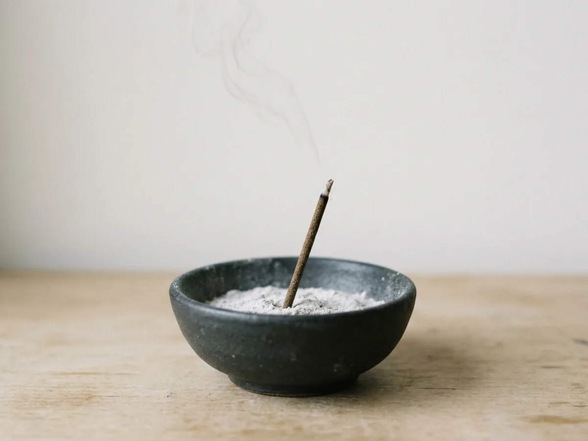 One short koh-grade incense stick set at a slight angle in a bed of fine white ash in a dark ceramic bowl