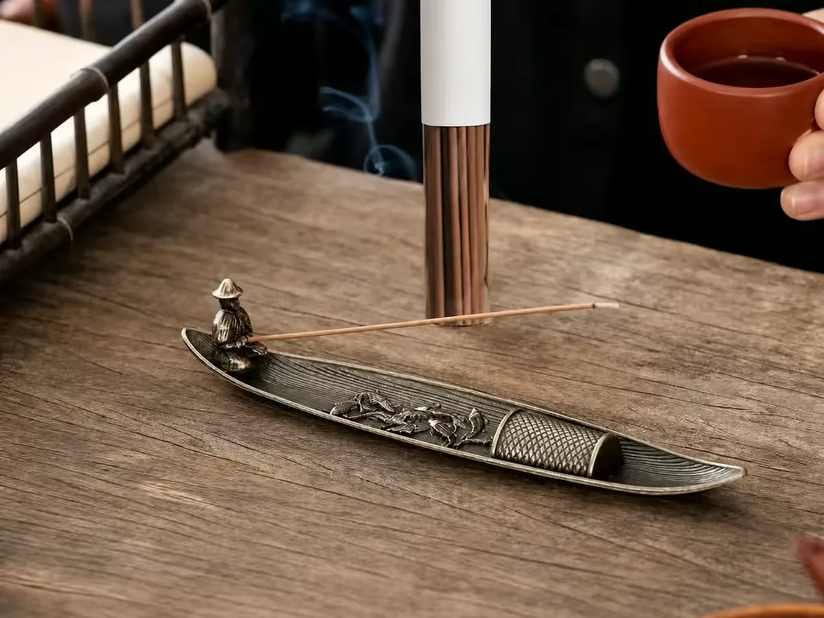 A lit incense stick on a boat holder at a tea table, a hand lifting a red tea cup beside it